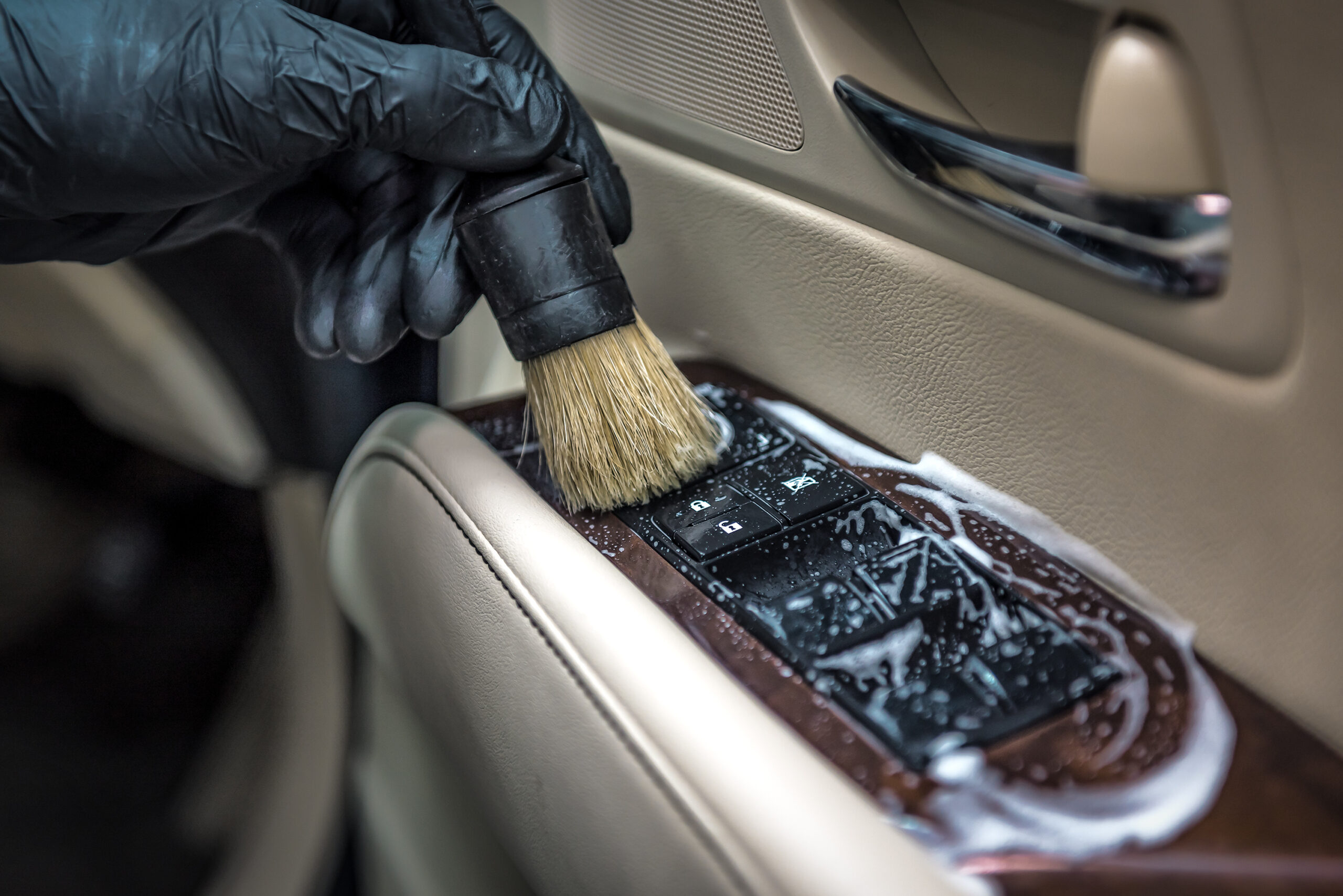 Detailing expert cleaning a car door panel with foam, focusing on unlock/lock and window buttons using a brush